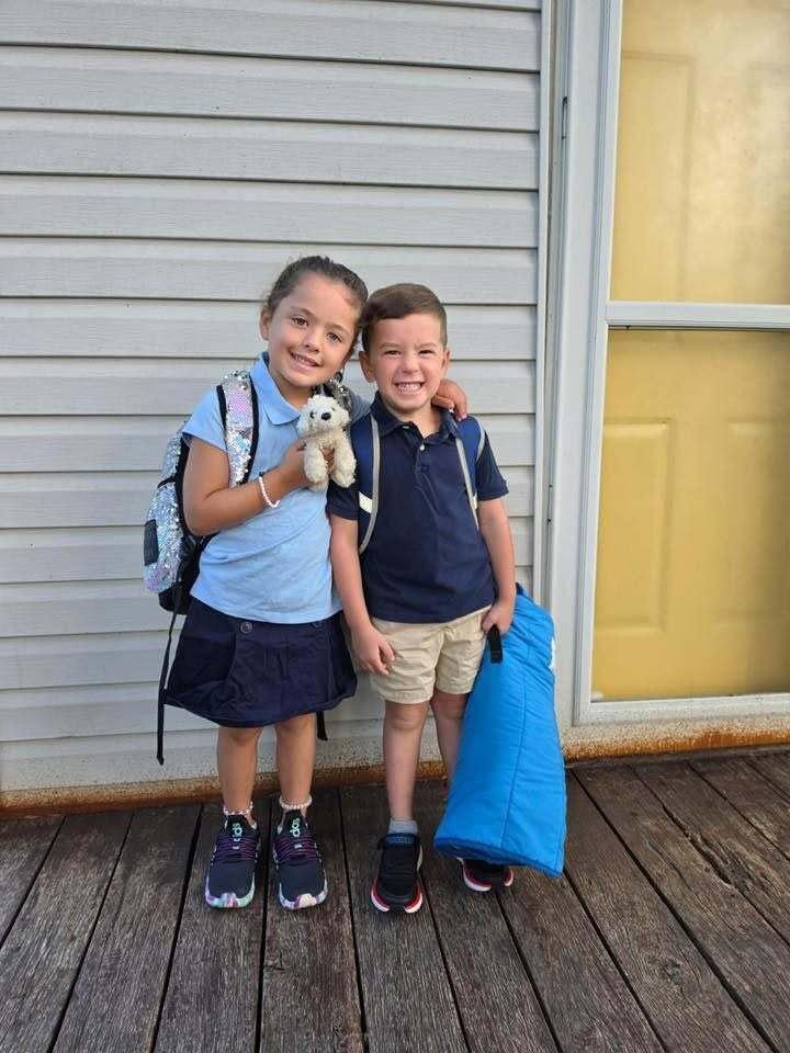 Two children stand outside a home and smile for a photo.