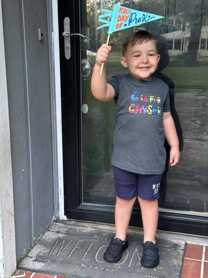 A child stands outside a home and poses for a photo. He holds up a first day of school flag.