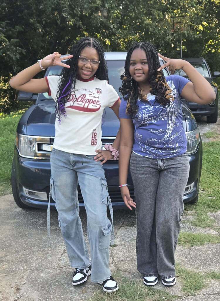 Two girls smile for a photo while standing outdoors in a driveway.