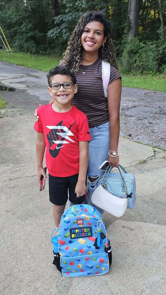 A boy and a girl smile for a photo outdoors in a driveway.