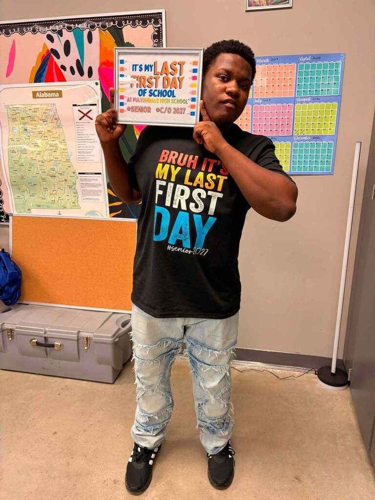 A Fultondale High School student poses for a photo in a room. The student holds up a sign that says "It's my last first day of school."