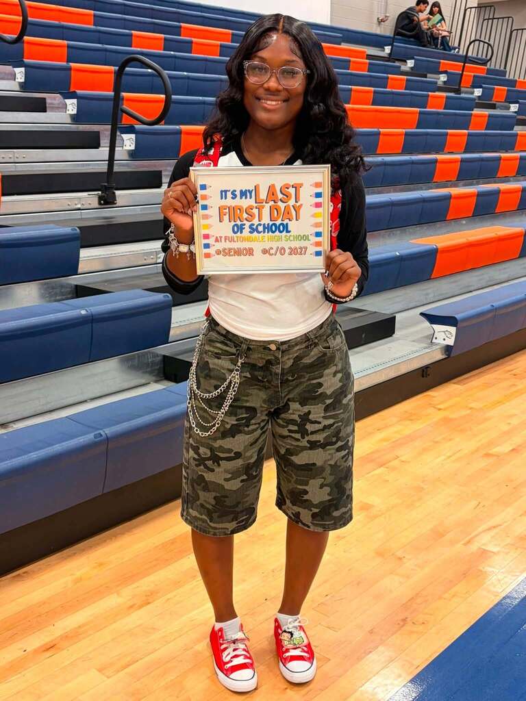 A Fultondale High School student smiles for a photo in the school gym. She holds up a sign that says "It's my last first day of school."