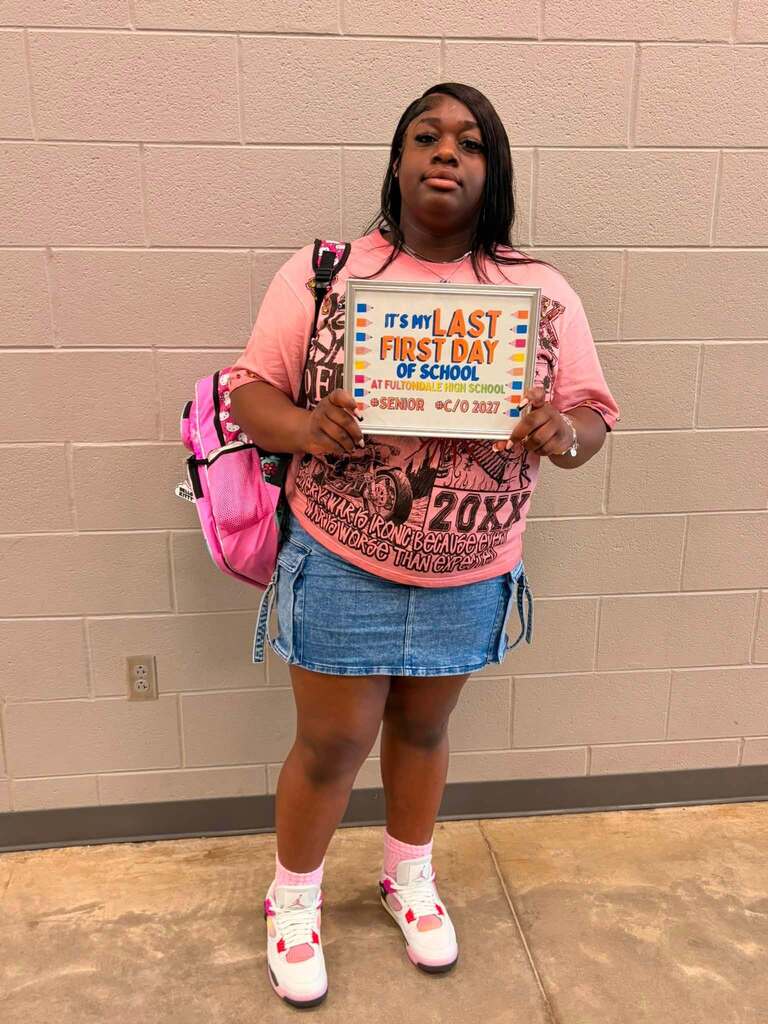 A Fultondale High School student poses for a photo in a school hallway. The student holds up a sign that says "It's my last first day of school."