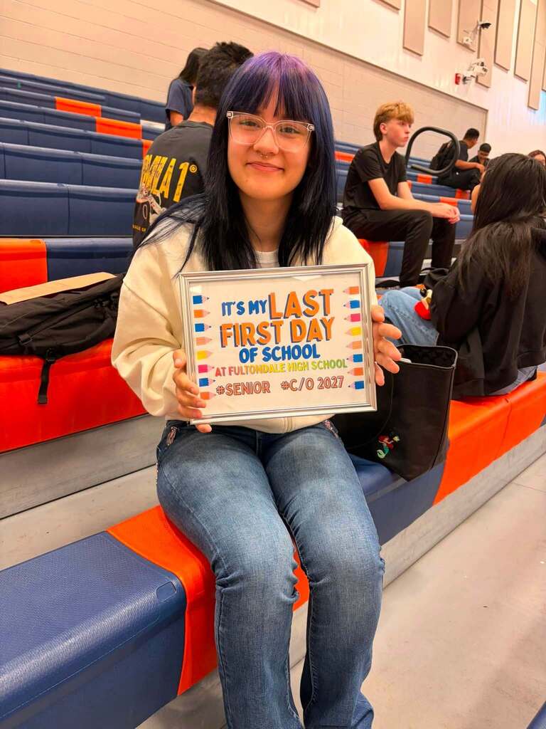 A Fultondale High School student smiles for a photo while sitting on bleachers and holds up a sign that says "It's my last first day of school."