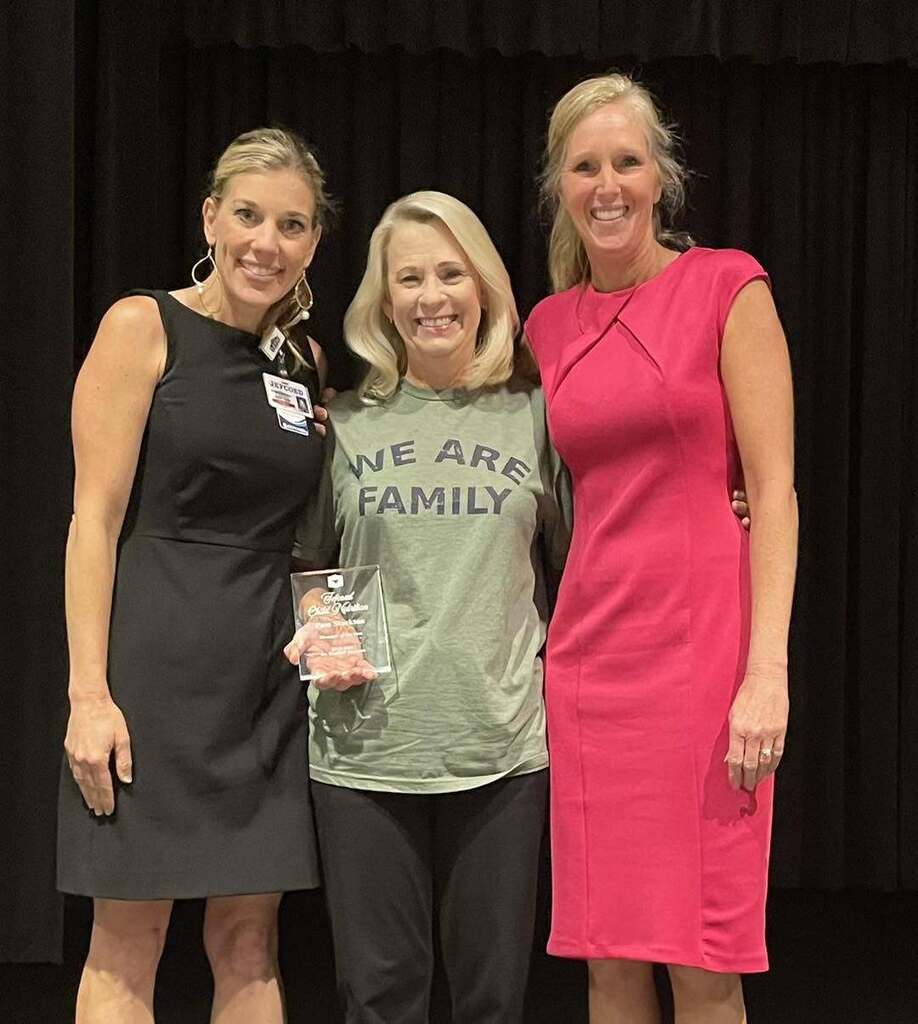 Three women stand together on a stage and pose for a photo. One of the women holds an award in her hand.