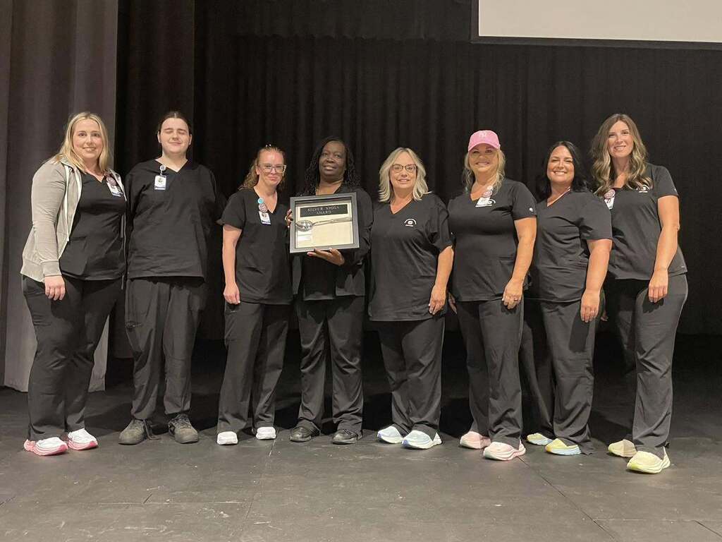 A group of women stand together on a stage and pose for a photo. One of them holds an award in her hands.