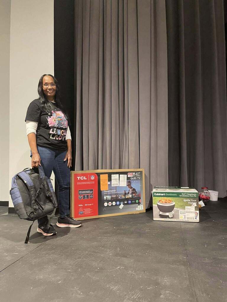 A woman smiles for a photo while standing on a stage. She stands next to a box with a TV in it and box with a fire pit in it.