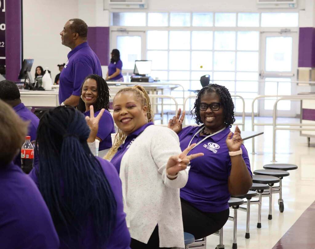 Educators sit at a lunchroom table and smile for a photo. They hold up peace signs with their hands.