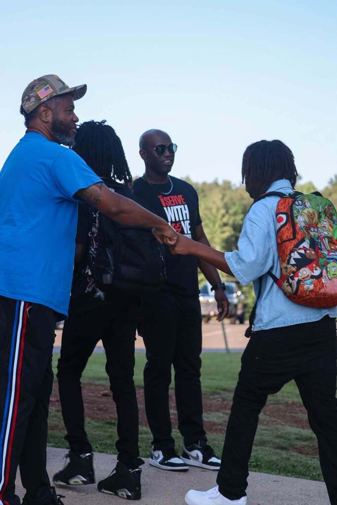 A group of men stand outdoors. One of the men gives a student a fist bump as the child walks by him.
