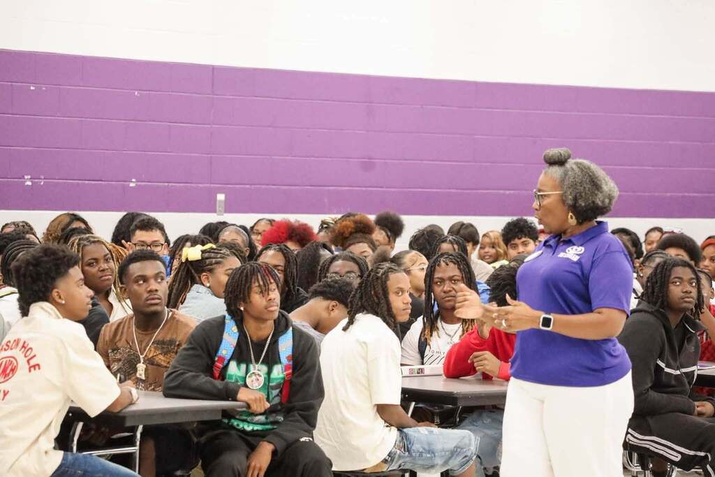 A woman speaks to a group of students as the students sit at lunchroom tables.
