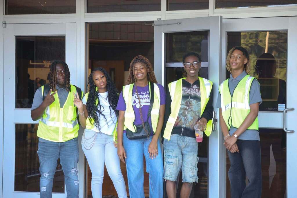 Students wearing yellow vests stand outside a school.