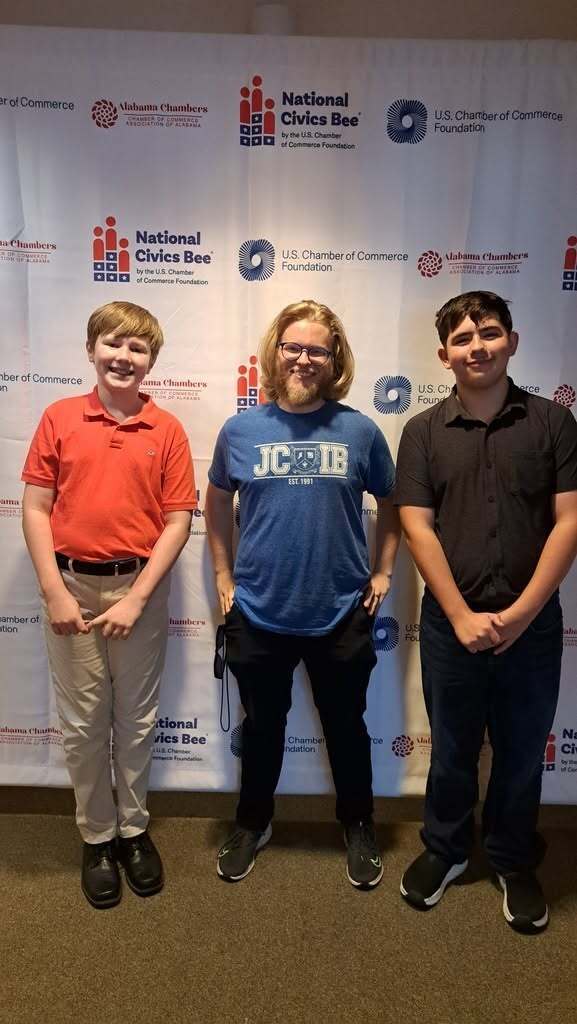 A teacher and two students stand together for a photo in front of a National Civics Bee photo backdrop.