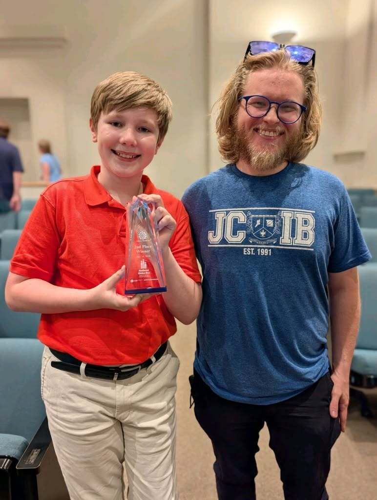 A student and a teacher stand together and smile for a photo. The student holds a glass award in his hands.