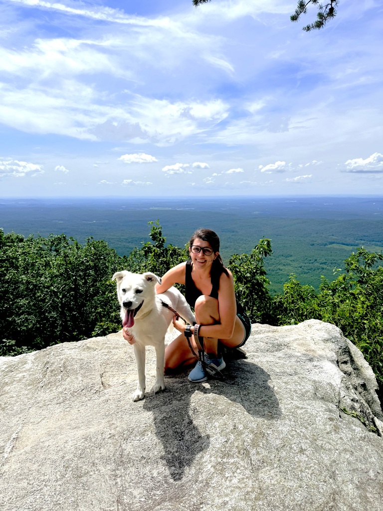 A woman poses for a photo with her dog while kneeling on a rock that looks over trees.