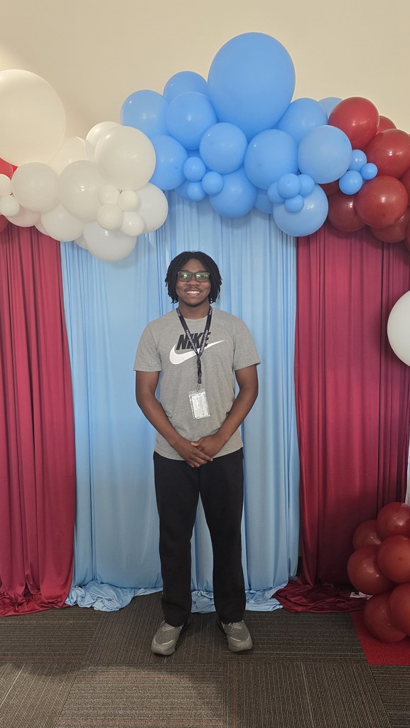 A student smiles for a photo while standing in front of a red and blue photo backdrop with balloons.