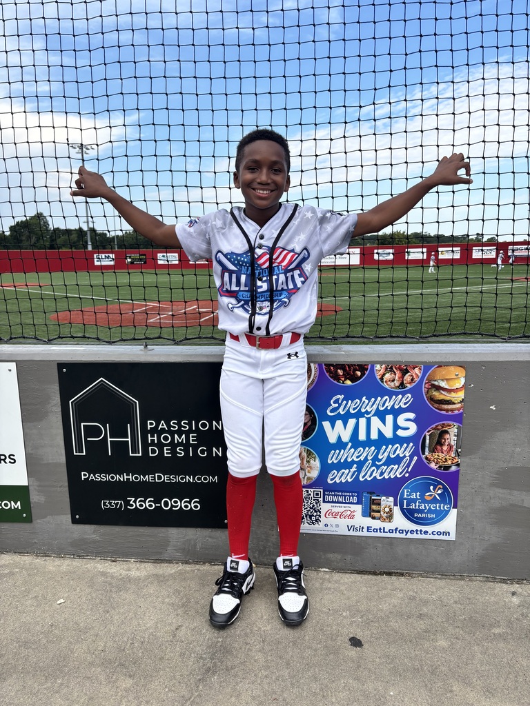 A child wearing a baseball uniform smiles for a photo in front of a baseball field.