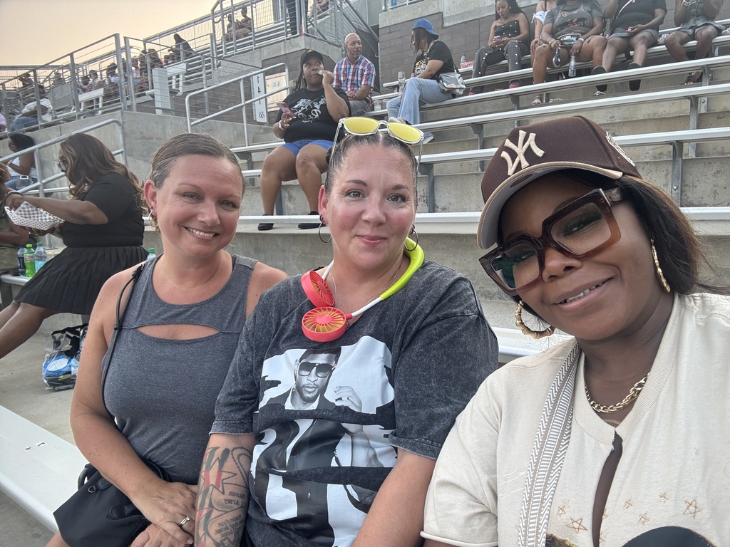 Three women smile for a photo while sitting in the bleachers of an open-air stadium.