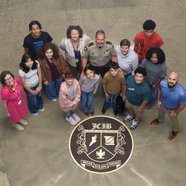 A group of 9 students, 4 staff and the SRO standing around the circular seal for JCIB featuring a coat of arms with a winged foot, a scroll with a feather quill, a flaming torch, and a banner reading "SECAN SCEARU FINDAN"