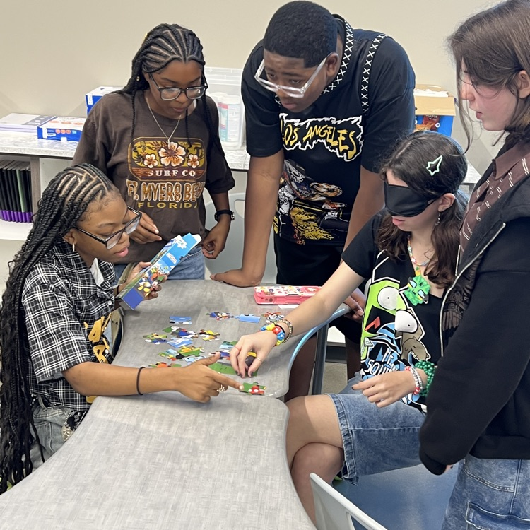 5 students working around a table. One student is blindfolded trying to put a jigsaw puzzle together while the others appear to be giving directions for how to do it.