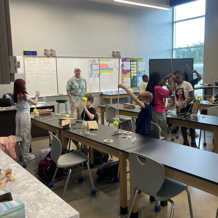 Sixth grade students around black lab tables working with a tennis ball, tape, q-tips, a cup , and wood to build the tallest tower. Mrs. Hume is standing at the front of the room watching.