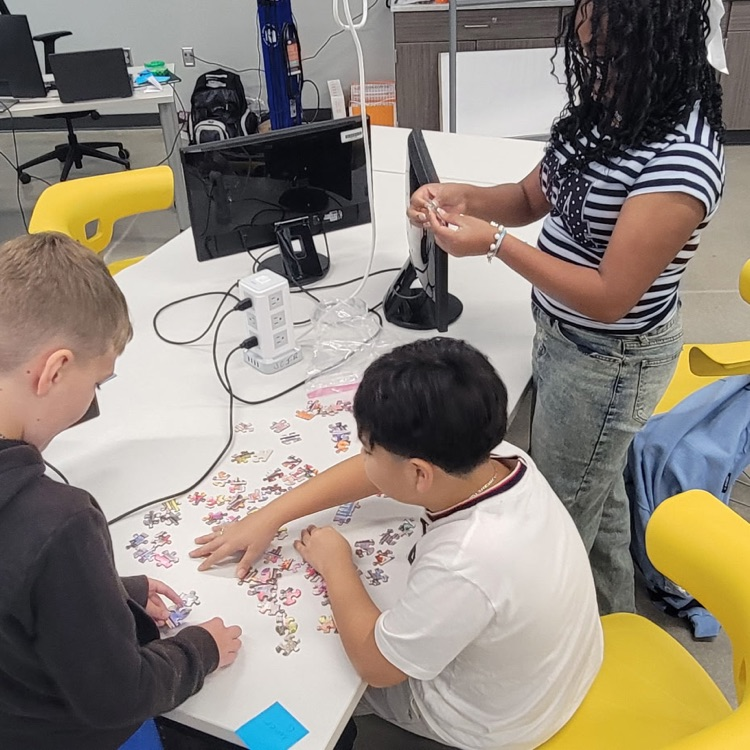 3 students working around a computer lab table putting pieces of a jigsaw together.