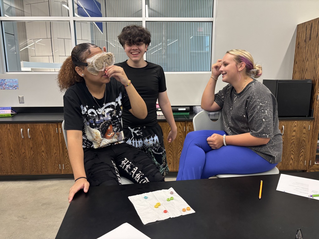 Three students sitting in stools around a lab table. One student is blind folders and taste testing fruit loops while her 2 peers watch.