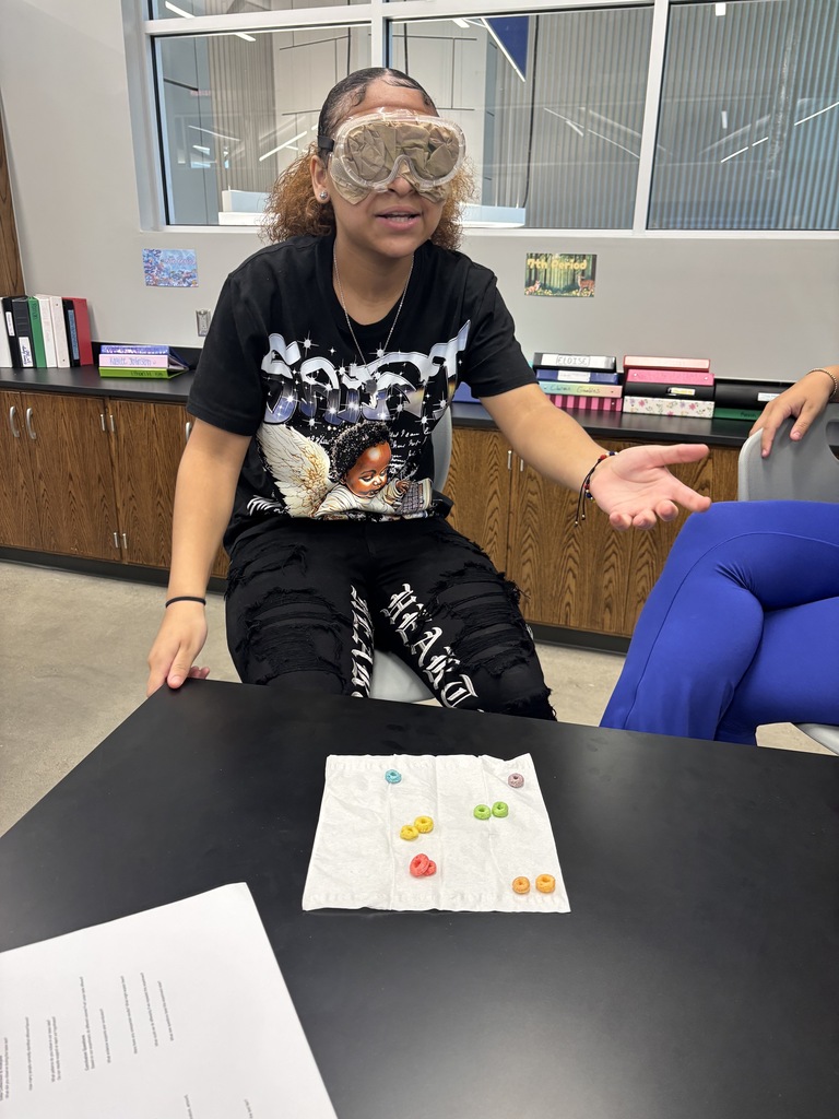 A student sitting in a stool at the lab science table with goggles that have been filled so she cannot see her surroundings. There are fruit loops on the paper towel in front of her.