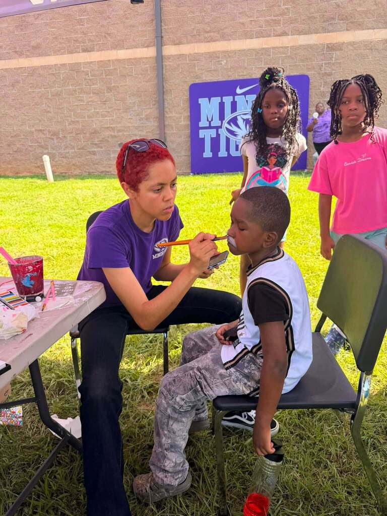 A person paints a child's face while sitting outdoors.