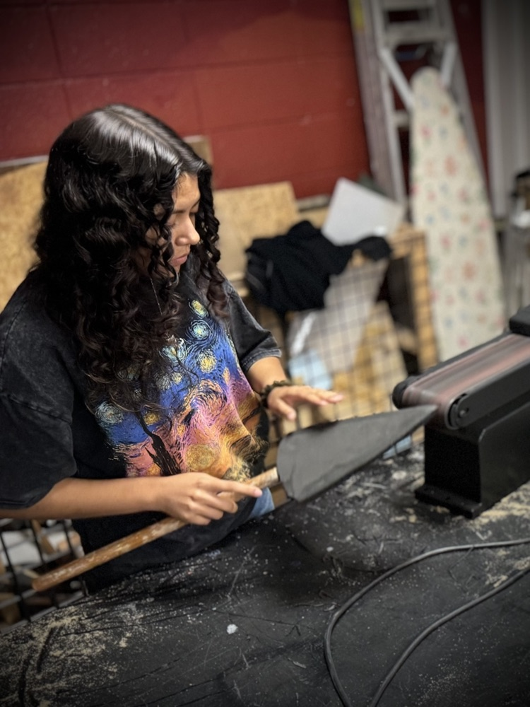 a student files the point of a foam spear on a belt sander