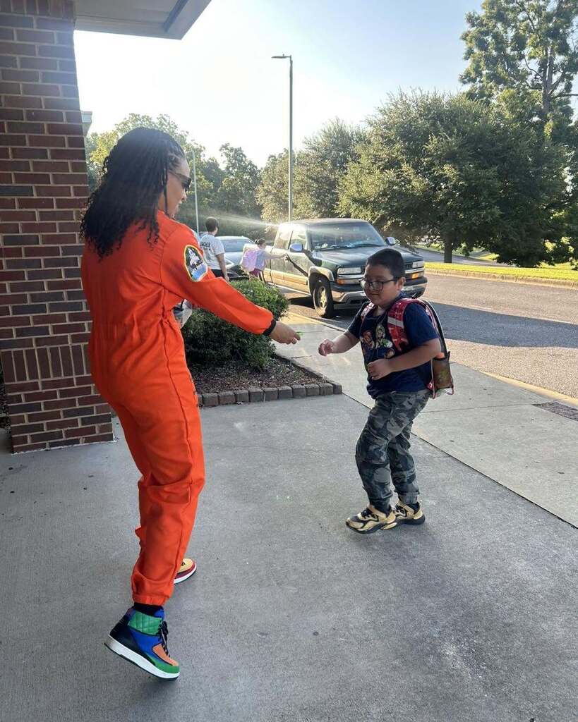 A woman wearing an astronaut costume hands a bracelet to a child outside a school.