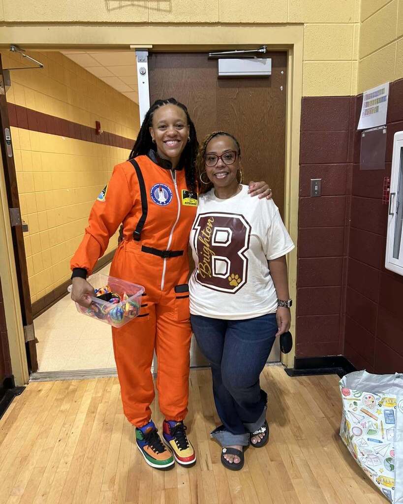 Two women stand together for a photo inside a school. One of the women wears an astronaut costume.