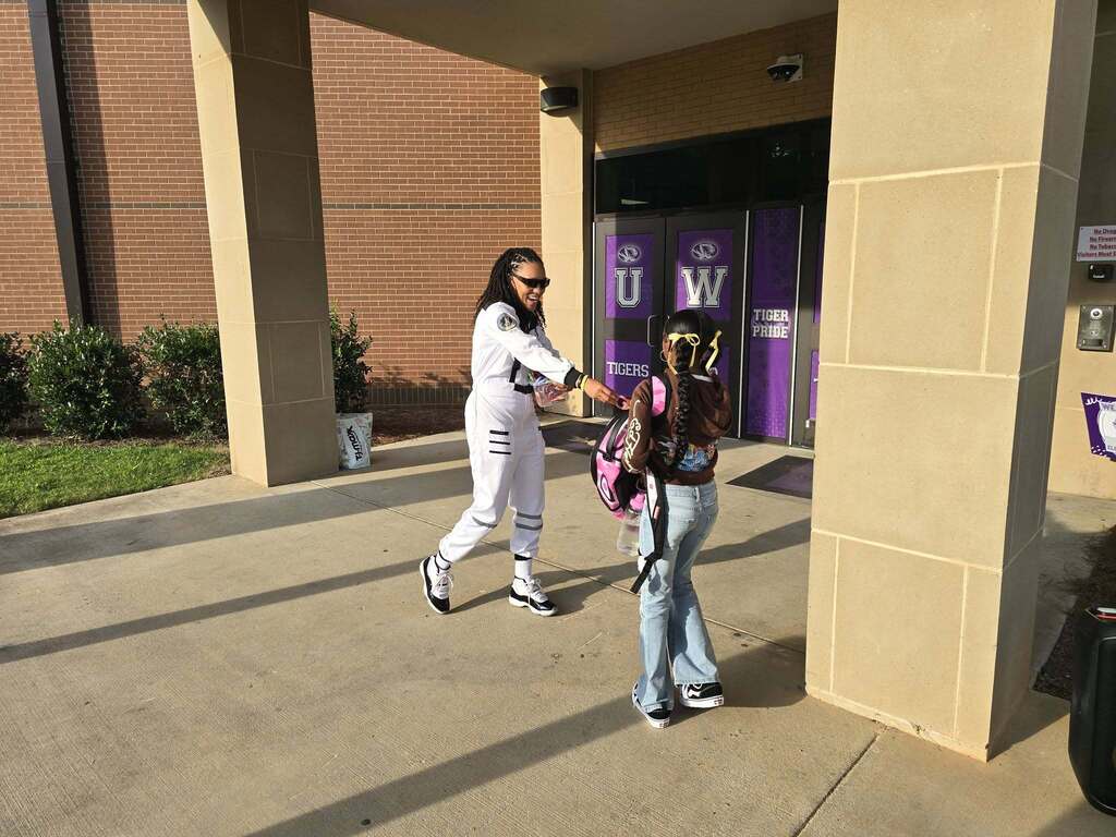 A woman smiles while handing a bracelet to a student outside a school. The woman wears an astronaut costume.