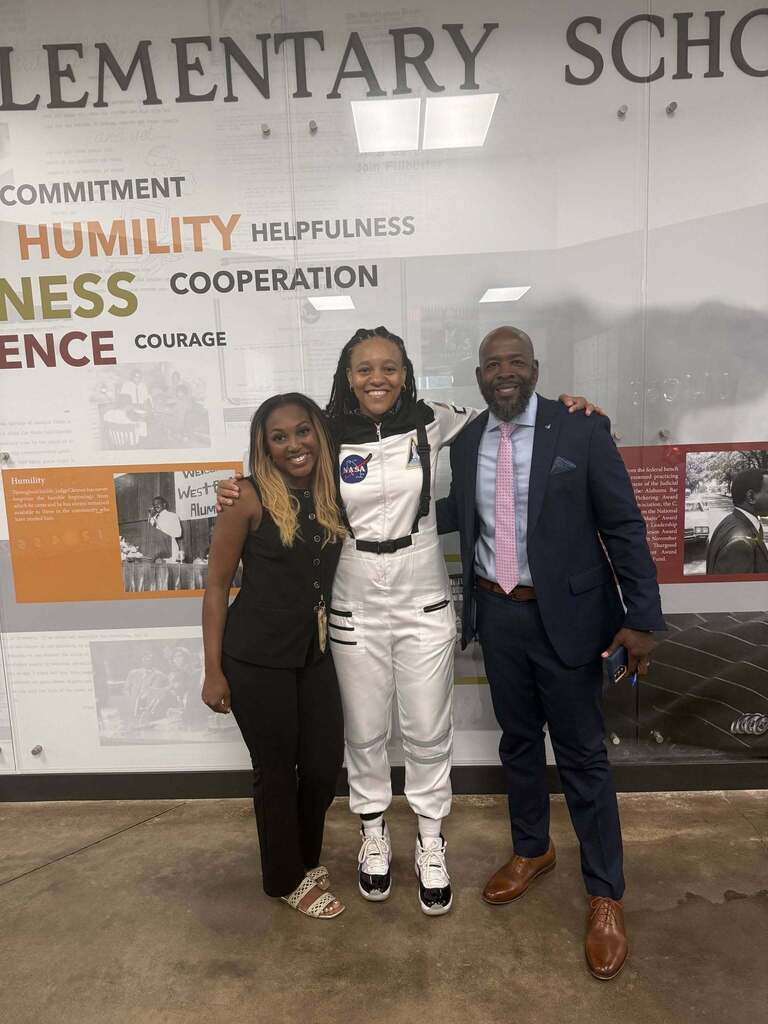 Three adults pose for a photo inside a school. One of them wears an astronaut costume.