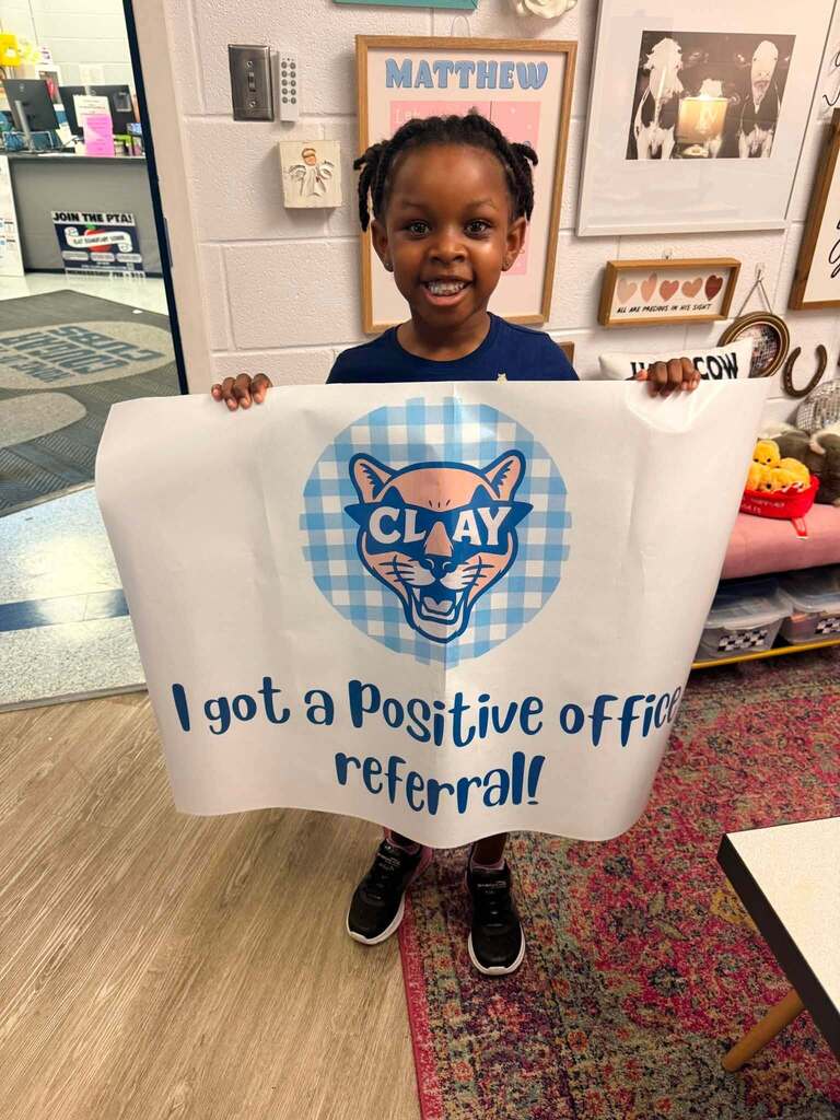 A child smiles and stands in a school office while holding a flag that says "I got a positive office referral!"