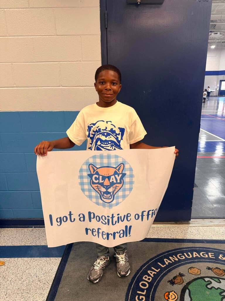 A child smiles and stands in a school hallway while holding a flag that says "I got a positive office referral!"