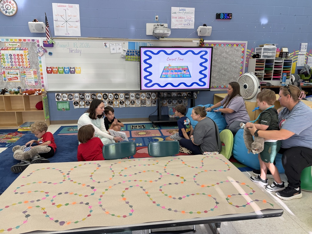 Picture of a classroom with 4 adults sitting on the floor or in chairs with 4 students sitting on the floor or standing. One adult in the middle is reading a story.