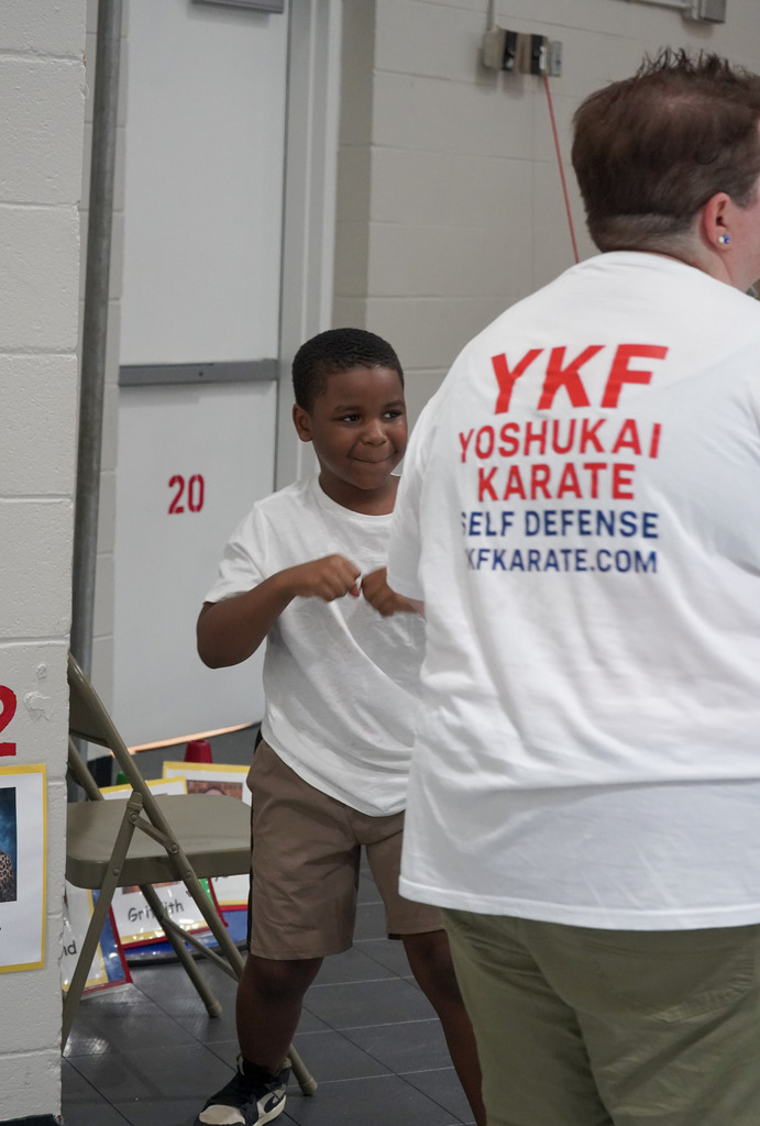 A child smiles while holding his fists up during a karate lesson in a school gym.