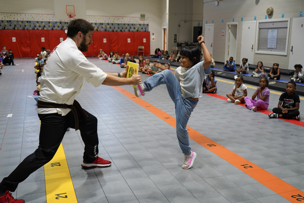 A student kicks a board being held by a karate instructor inside a school gym.