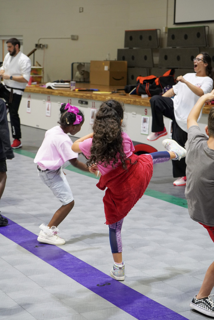 Children kick their legs in the air during a karate lesson inside a school gym.