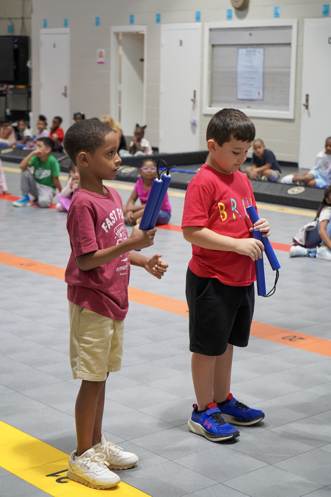Two children stand in a school gym and hold nunchucks in their hands.