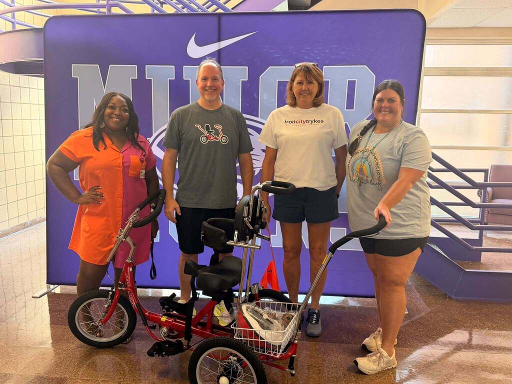 Four adults smile for a photo in front of a Minor High School photo backdrop. A therapeutic tricycle sits on the floor in front of them.