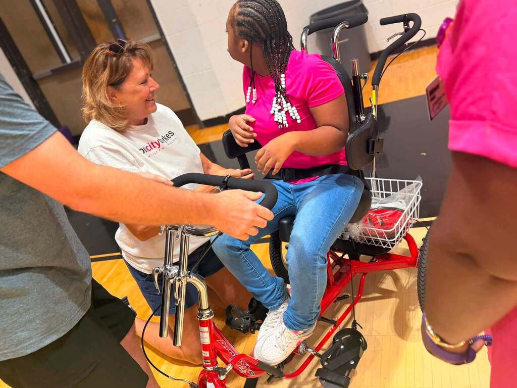 A woman smiles while kneeling next to a student sitting on a therapeutic tricycle.