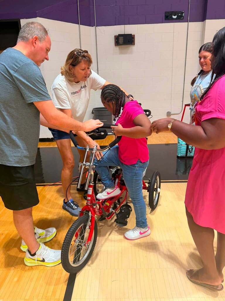Adults help a student climb onto a therapeutic tricycle inside a school gym.