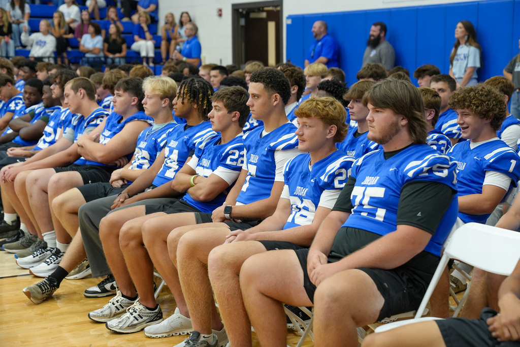 Football players sit in chairs inside a school gym.