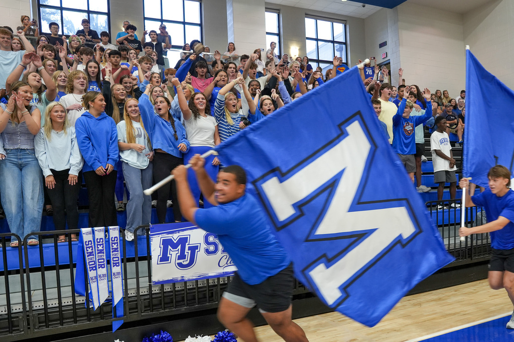Students stand in bleachers and cheer while other students run across the gym floor holding large flags.