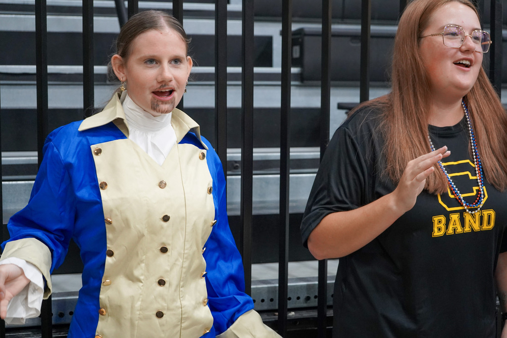 Students stand in a school gym during a pep rally. One of the students wears a Hamilton costume.
