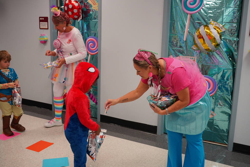 Adults dressed in cnady land themed costumes in front of their doors decorated with candy. Student in spiderman costume is receiving candy. 