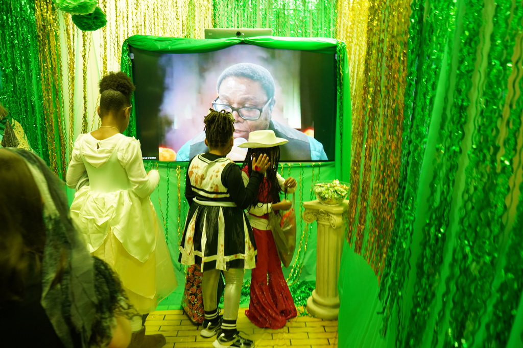 Students standing in a room decorated green to look like the Oz from The Wizard of Oz. Someone is projecting themselves on the tv screen as "The Oz". The students are getting candy from a bowl in front of the screen. 