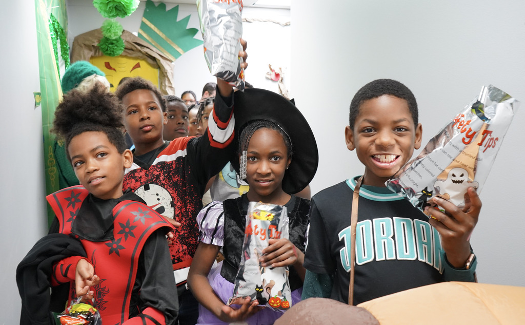 Children wearing Halloween costumes smile for a photo while standing inside a hallway.