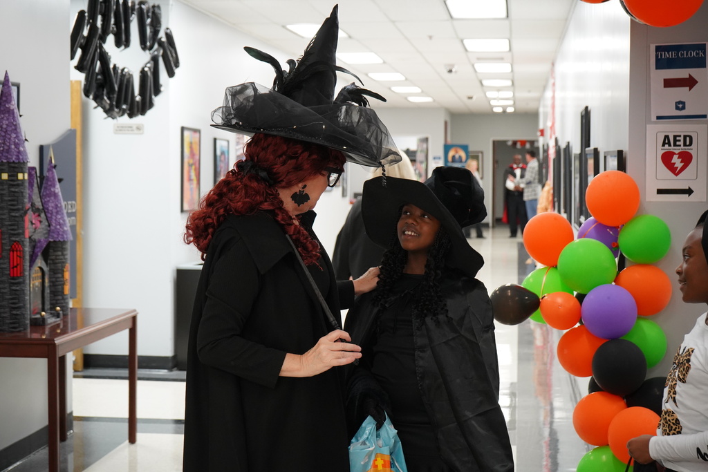 A woman and a student wearing witch costumes smile and speak to each other while standing inside a building.
